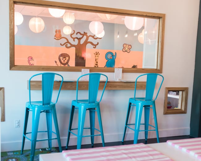 The image shows a brightly lit room featuring a seating area with turquoise bar stools and a large window overlooking a colorful play area.