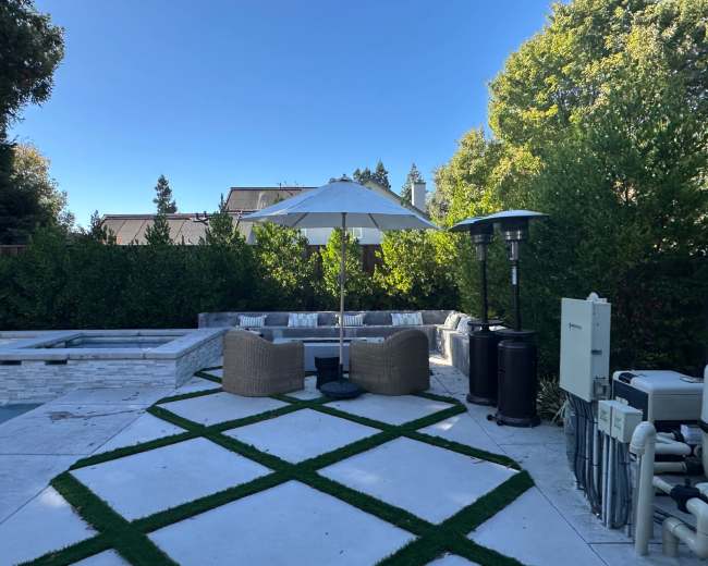 The image shows a patio area with two wicker chairs under a large white umbrella, beside a pool and a heated patio heater, surrounded by greenery.