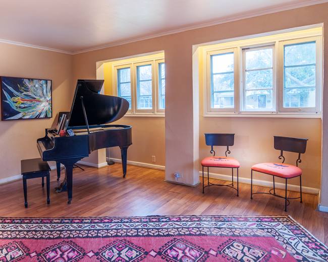 A room featuring a grand piano, two chairs, and framed artwork on the walls, with windows allowing natural light to enter.