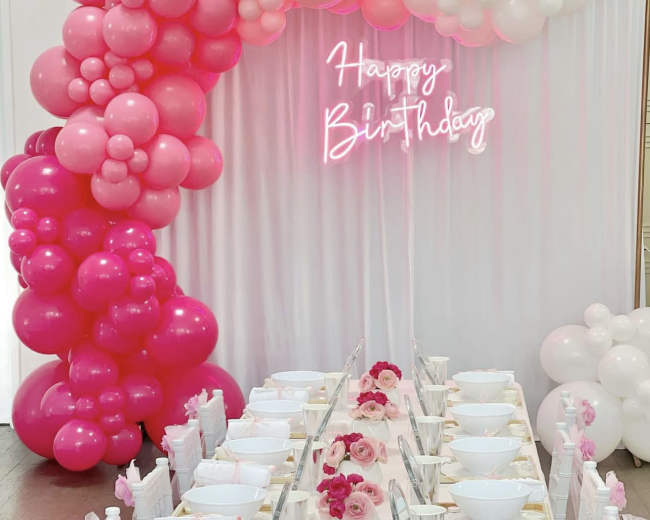 A table is set for a birthday celebration, with a decorative balloon arch in shades of pink and white and a neon "Happy Birthday" sign.