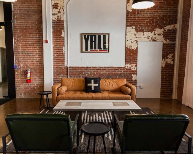 The image shows a modern lounge area featuring a leather couch, two green chairs, a coffee table, and exposed brick walls.