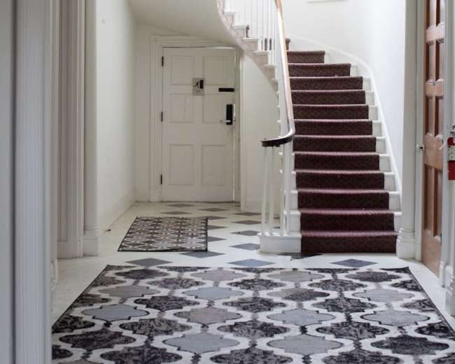 The image shows a spacious hallway featuring a curved staircase, a chandelier, decorative rugs, and a door leading outside.