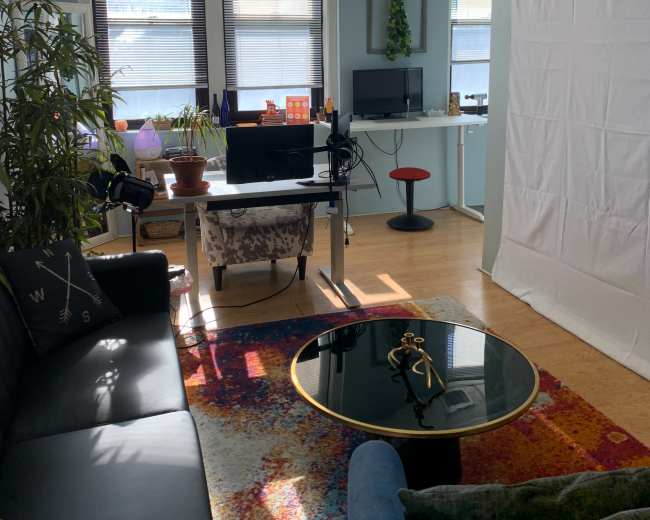 A well-lit office space with a couch, a coffee table, a desk with a monitor, and a backdrop setup in one corner.