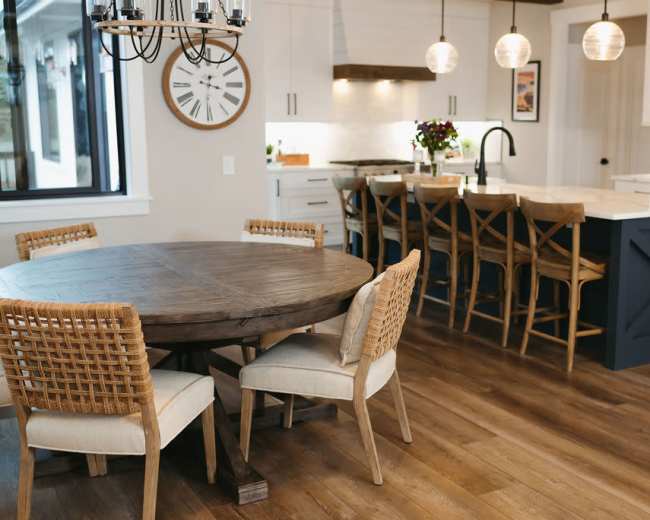 A round wooden dining table surrounded by chairs is set in a bright kitchen with an island and bar stools, featuring modern light fixtures and large windows.