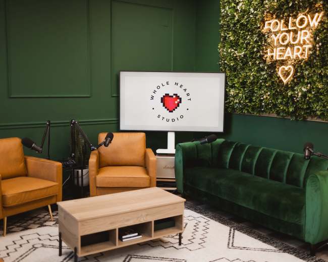 The image shows a cozy studio space with green walls, a logo on a screen, two armchairs, a sofa, and decorative plants alongside a neon sign that reads "FOLLOW YOUR HEART."
