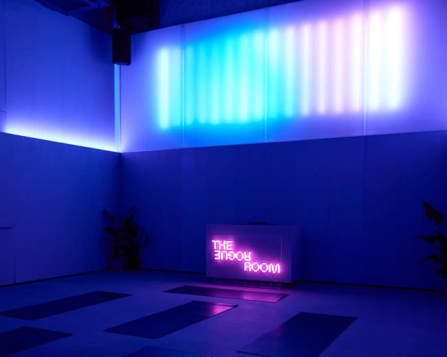 The image shows a dimly lit yoga studio with blue and purple ambient lighting and several yoga mats arranged on the floor.