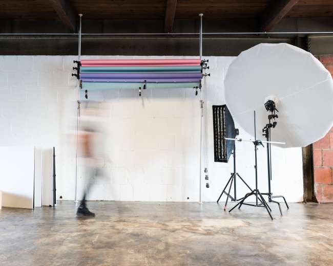 A blurred figure walks past a photography setup featuring backdrop rolls and studio lighting in an industrial space.