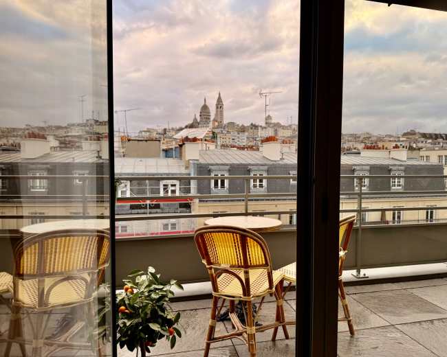 The image shows a balcony with two wicker chairs facing a cityscape featuring prominent buildings under a cloudy sky.