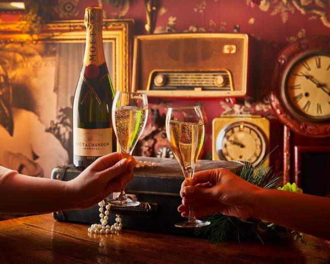 Two hands clink champagne glasses over a wooden table adorned with a vintage suitcase, surrounded by old photographs and retro decor.