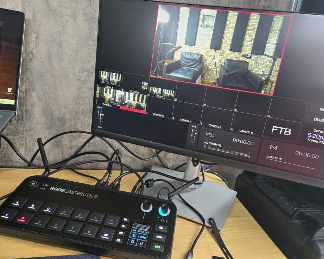 A computer monitor displays a video production interface featuring a preview of a studio setup with two empty chairs and a control panel for managing media and recording.