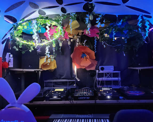 A DJ setup within a decorated booth featuring colorful flowers and greenery, with sound equipment and a keyboard visible in the foreground.