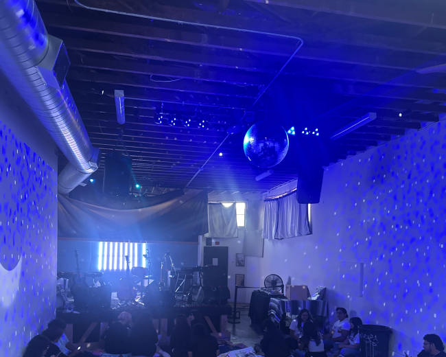 A group of people is sitting on the floor of a dimly lit room with colorful lighting and musical equipment set up at the front.