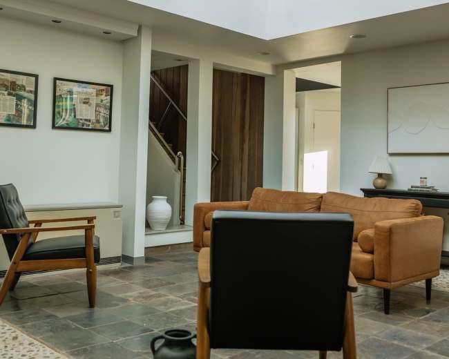 The image shows a minimalist living room with a brown sofa, a black chair, and a large window allowing natural light to enter.