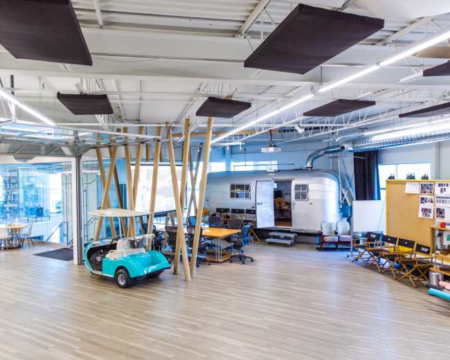 The image shows an open office space featuring a retro camper, tables, chairs, and a golf cart, with large windows allowing natural light to enter.