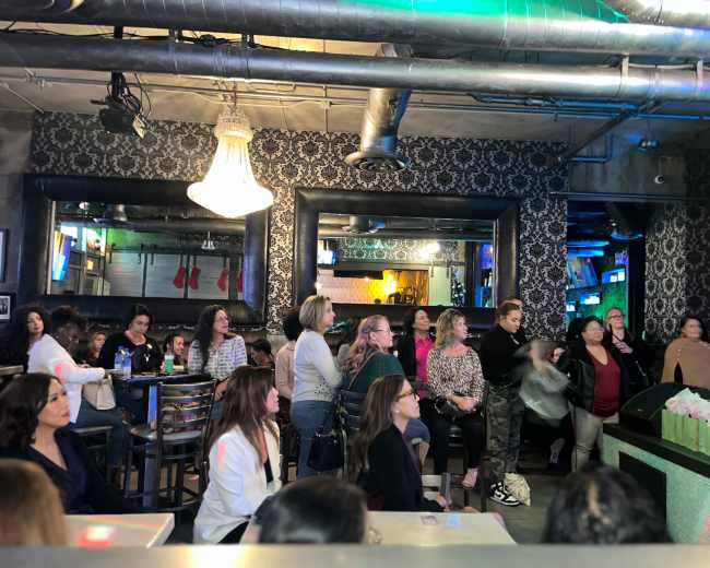 A diverse group of women is gathered in a dimly lit bar, watching a performance with several mirrors and decorations in the background.