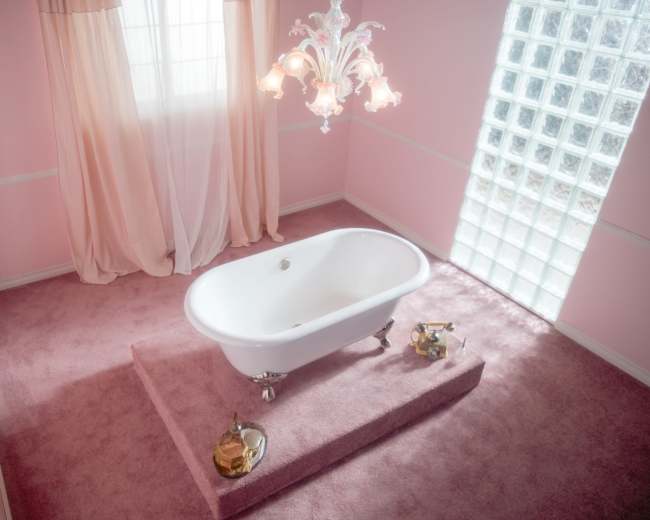 A white freestanding bathtub is positioned on a raised platform in a softly lit pink bathroom with a chandelier and a window featuring glass blocks.