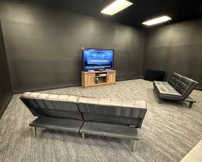 The image shows a sparsely furnished room with black walls, featuring a television on a wooden stand, a brown leather couch, and two additional seating options against the walls.