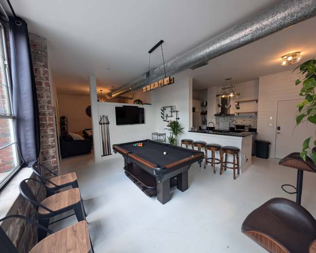 The image shows a spacious interior featuring a billiard table in the center, with seating areas, a kitchen counter, and large windows providing natural light.