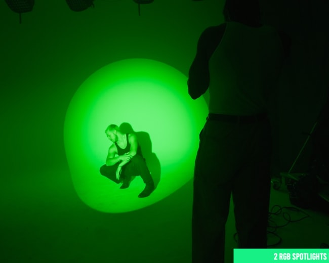 A person crouches in a green-lit studio while another figures are positioned behind a camera.