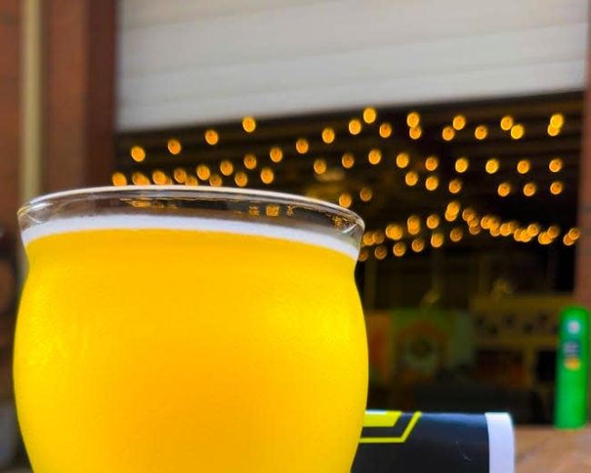 A glass of yellow beer sits on a wooden table in front of a branded coaster featuring a cartoon character, with a backdrop of softly lit string lights.