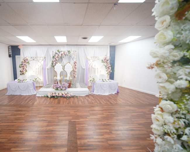 The image shows a decorated event space featuring a stage with floral arrangements, a central backdrop, and tables set up for a formal occasion.