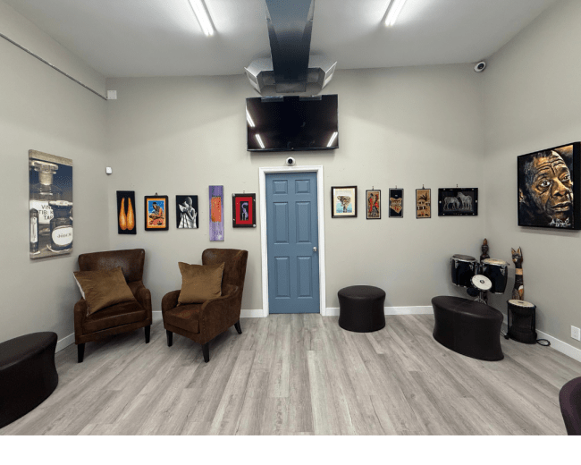 The image shows a modern room with two brown armchairs, a variety of framed artworks on the walls, a television mounted above a blue door, and a small drum set in the corner.