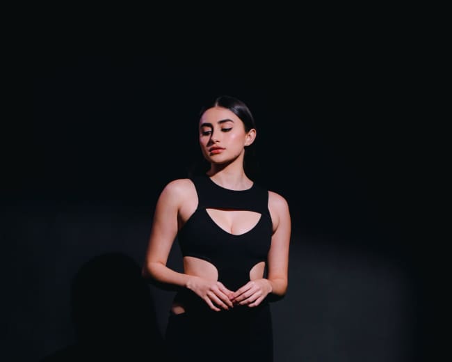 A woman stands against a dark background, wearing a black cut-out dress and high heels.