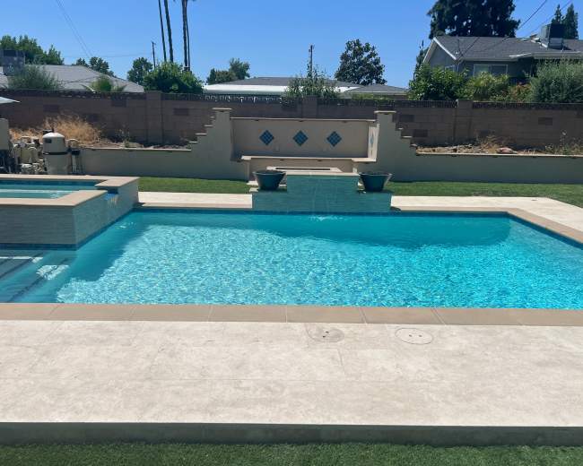 The image shows a clear swimming pool with a spa section, surrounded by a stone patio and a landscaped area featuring grass and decorative plants.