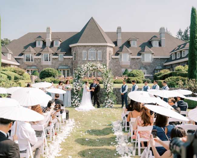 A couple stands under a floral arch during their wedding ceremony in front of a large, elegant mansion, surrounded by guests seated on white chairs.