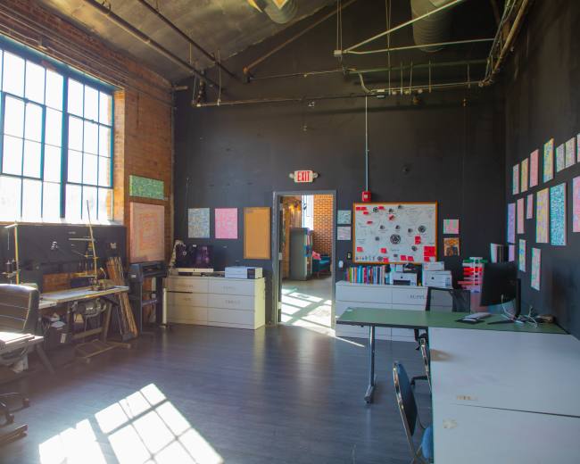 The image shows a spacious office with large windows, featuring several desks, chairs, and colorful artwork on the walls.