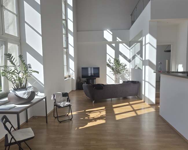 The image shows a spacious, modern living area with large windows casting diagonal shadows on the wooden floor.
