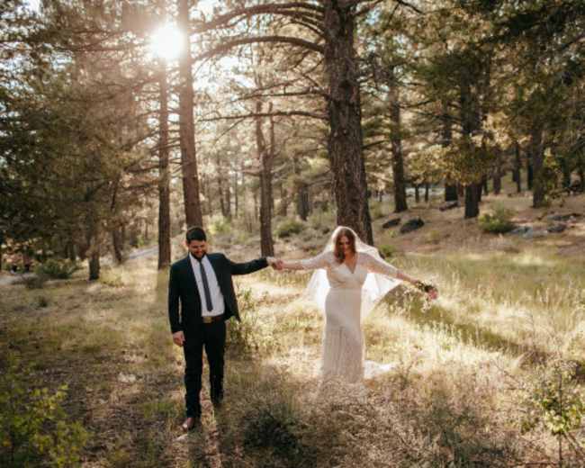 A couple walks hand in hand through a sunlit forest, with the woman wearing a wedding gown and the man in a suit.