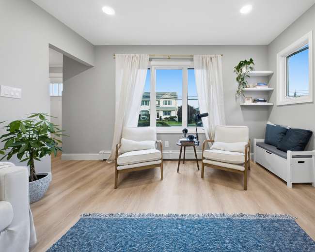 The image shows a modern living room featuring two armchairs, a small table, a window with natural light, and a plant near the corner.