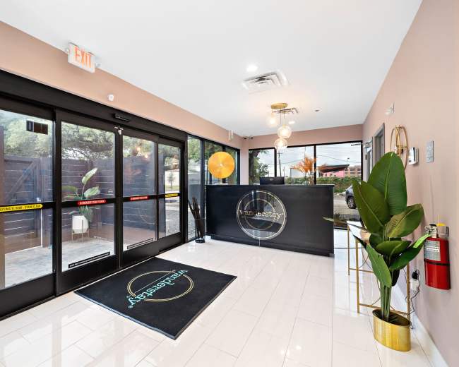 The image shows a modern reception area with large glass doors, a sleek front desk, and stylish indoor plants, all featuring neutral tones and bright lighting.