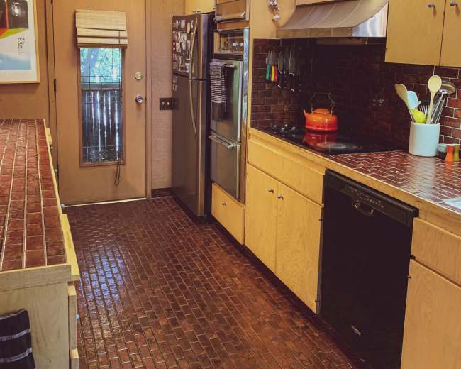 The image shows a modern kitchen featuring wooden cabinetry, a stainless steel refrigerator, and a tiled floor with a red brick pattern.