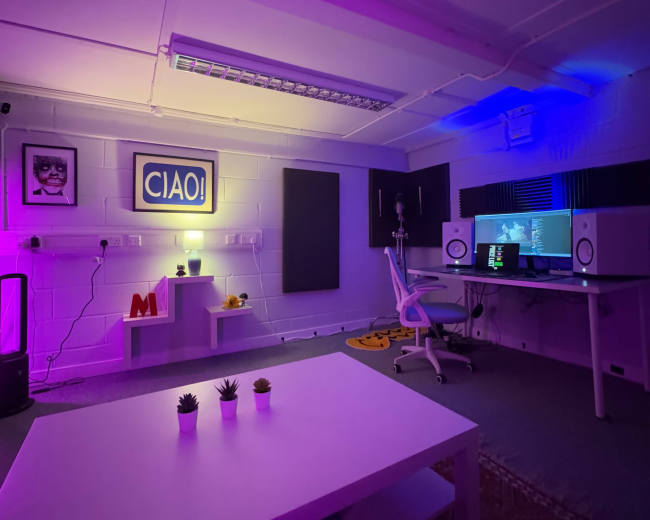 A studio room features a white table, a comfortable chair, colorful LED lighting, and workstations with monitors and sound equipment.