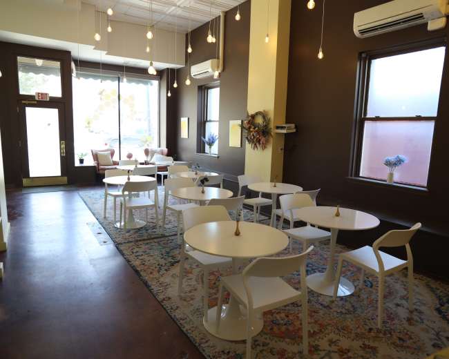 The image shows a bright café interior with several white tables and chairs arranged on a colorful rug, large windows letting in natural light, and light fixtures hanging from the ceiling.