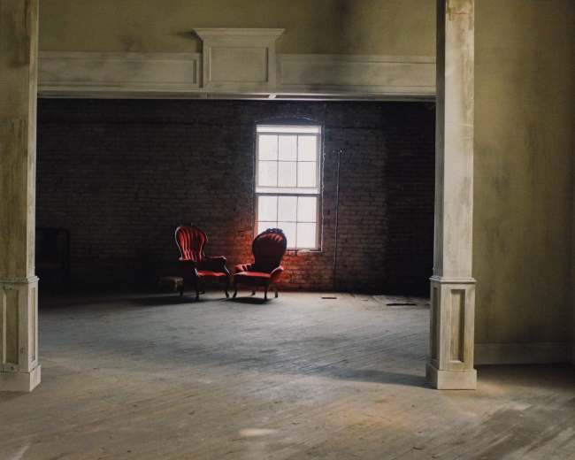 A pair of red chairs is positioned in a dimly lit room with exposed brick walls and a large window.