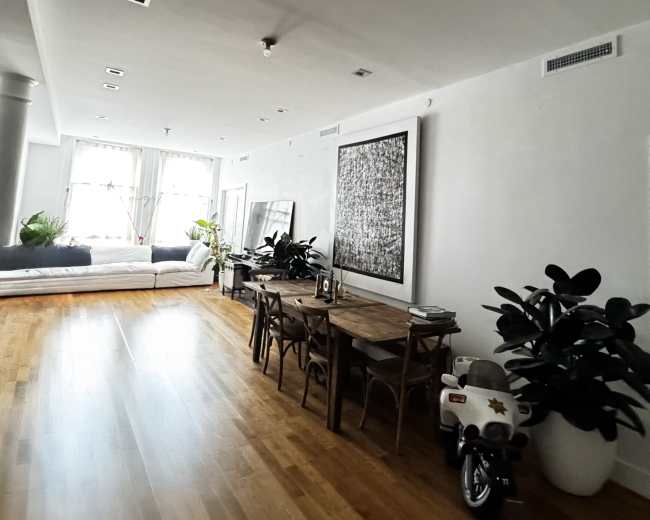 The image shows a bright, modern living space with a dining table, chairs, and plants, beside a bed in a separate area.