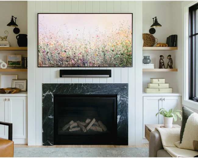 The image shows a modern living room featuring a fireplace with a dark stone surround, a large TV mounted above it displaying a floral artwork, and shelves on either side filled with decorative items.