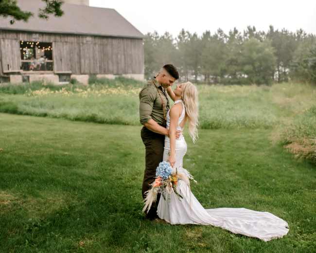 A couple stands in an open field, embracing each other with a rustic barn and trees in the background.