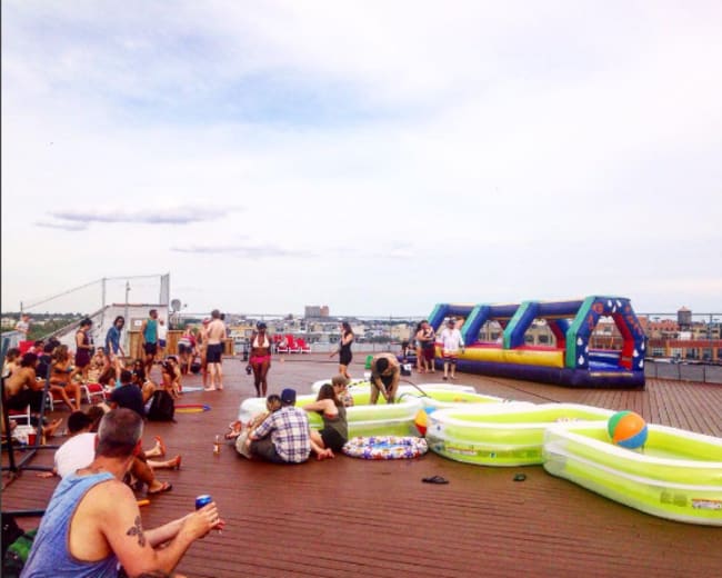 A rooftop party features people lounging around inflatable pools and a bounce house with a scenic waterfront background.