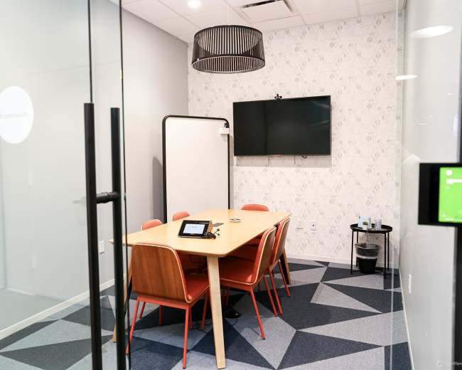 The image shows a modern meeting room with a wooden table, four orange chairs, a flat-screen TV mounted on the wall, and a whiteboard.