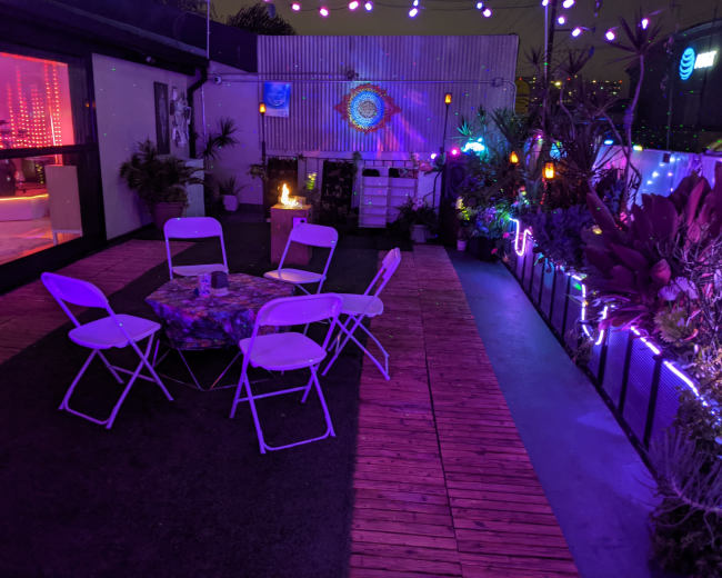 A patio area features a circular arrangement of white folding chairs around a colorful table, surrounded by plants and illuminated by colorful string lights.