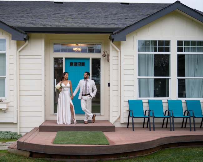 A couple dressed in formal attire is walking hand in hand on the porch of a house with teal accents and a row of blue chairs.