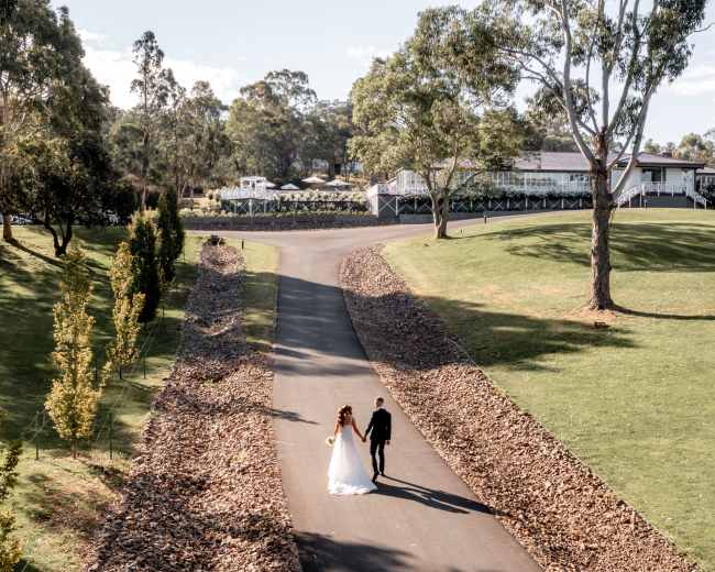 A couple walks hand in hand down a tree-lined path leading to a building on a hillside.
