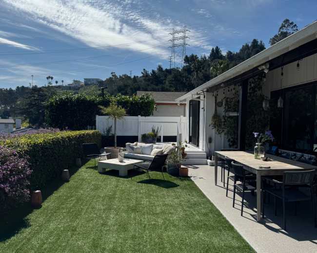 The image shows a landscaped outdoor patio area with artificial grass, a seating arrangement, and a dining table overlooking a hillside.