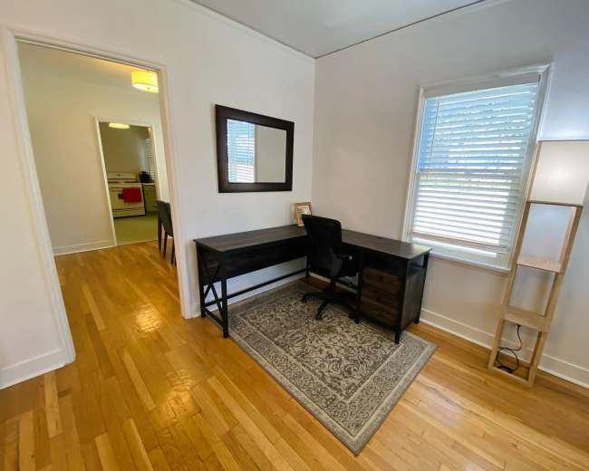 A small office space features a dark wooden desk with a chair, a mirror on the wall, and a floor lamp beside a window, with a view of an adjacent room.