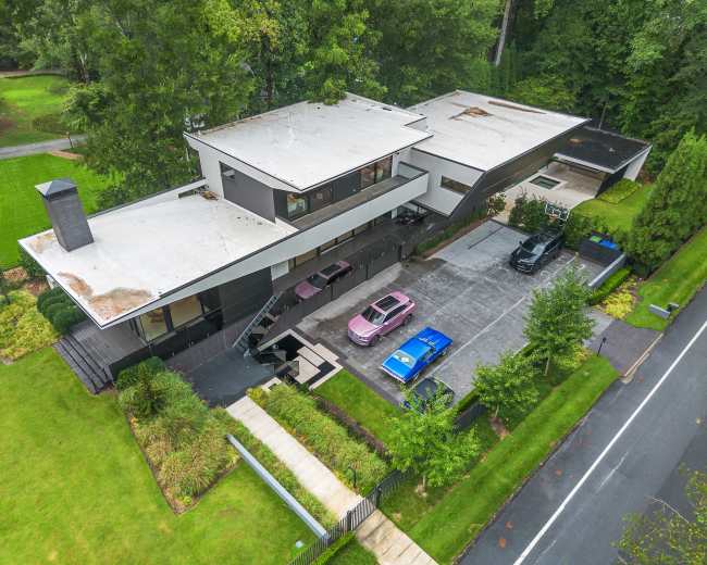 The image shows a modern house with flat roofs, situated on a lawn and featuring several parked cars in a driveway.