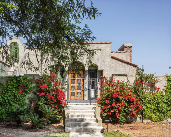 A stucco house with wooden doors is surrounded by vibrant flowering plants and has stairs leading to the entrance.